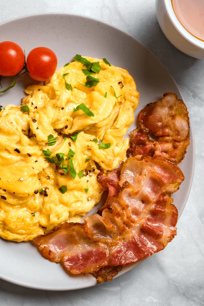 A plate of scrambled eggs garnished with chopped herbs, two strips of crispy bacon, and two cherry tomatoes, prepared using an air fryer.