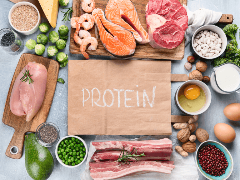 A variety of high-protein foods including salmon fillets, shrimp, red meat, chicken breast, cheese, eggs, milk, white beans, Brussels sprouts, broccoli, peas, avocado, nuts, seeds, and pomegranate seeds.