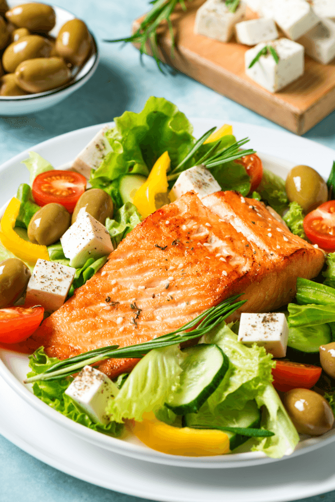 A plate of cooked salmon on a bed of fresh salad including lettuce, cherry tomatoes, cucumber slices, yellow bell pepper strips, green olives, and feta cheese cubes, garnished with rosemary.