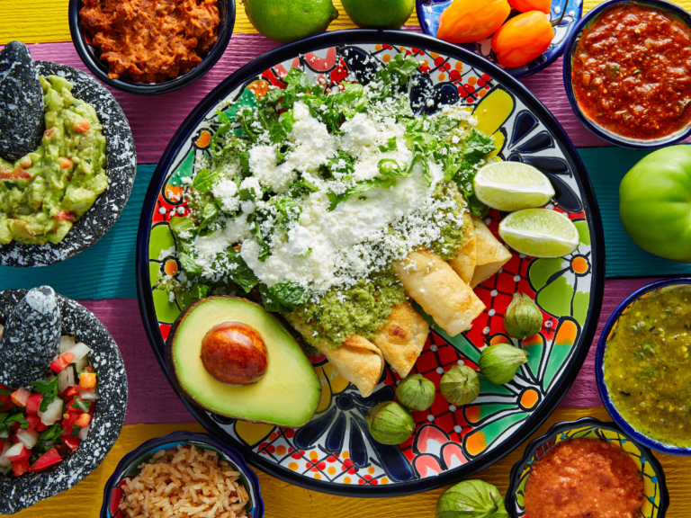 A colorful Mexican meal spread featuring enchiladas with green sauce, crumbled cheese, fresh greens, guacamole, salsa, pico de gallo, rice, and lime wedges.