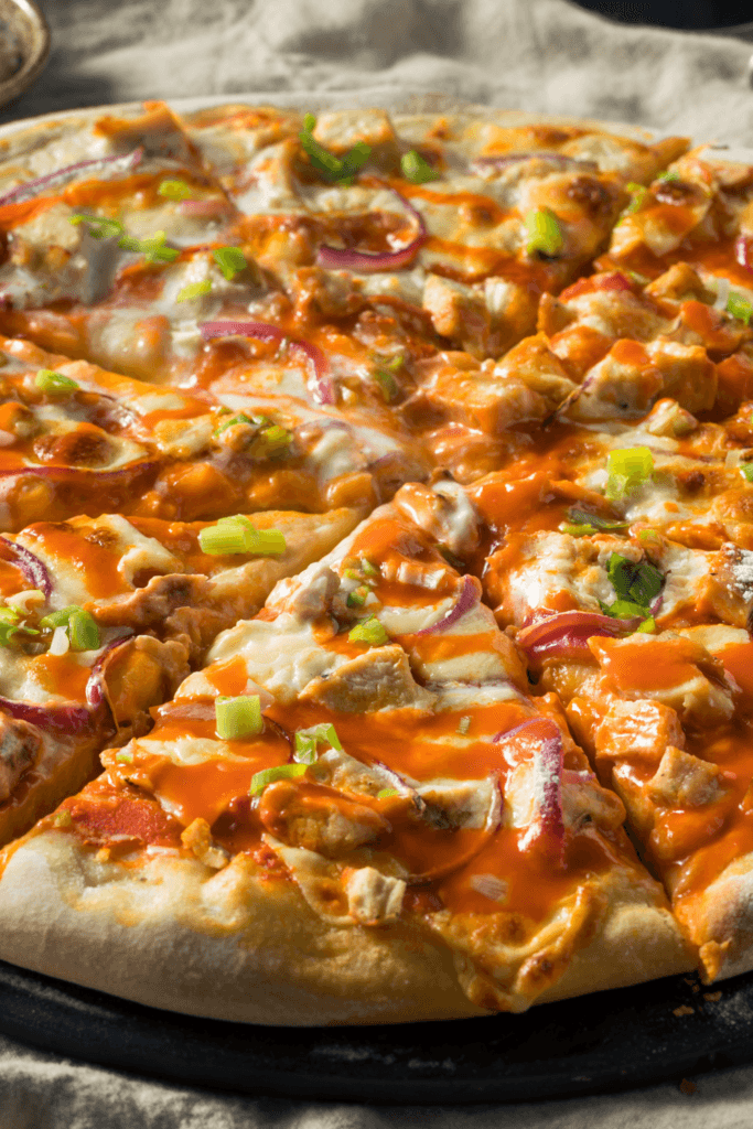 A close-up view of a Buffalo Chicken pizza topped with pieces of chicken, red onion slices, green onions, melted cheese, and a generous amount of buffalo sauce on a thick, slightly browned crust.