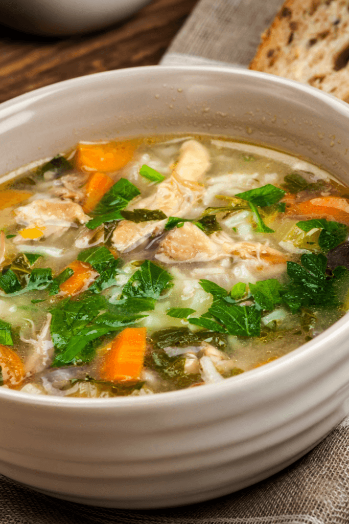 A bowl of chicken fajita soup with pieces of chicken, chopped carrots, and green leafy herbs, served with a piece of crusty bread in the background.