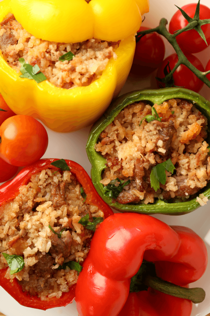 Three stuffed bell peppers of yellow, green, and red, filled with a mixture of rice, ground meat, and herbs, surrounded by small red tomatoes still attached to their vine.