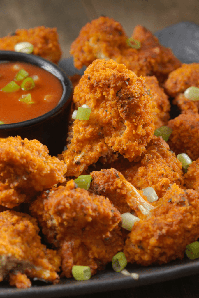 A plate of crispy breaded cauliflower florets garnished with chopped green onions, served with a small black bowl of red dipping sauce.