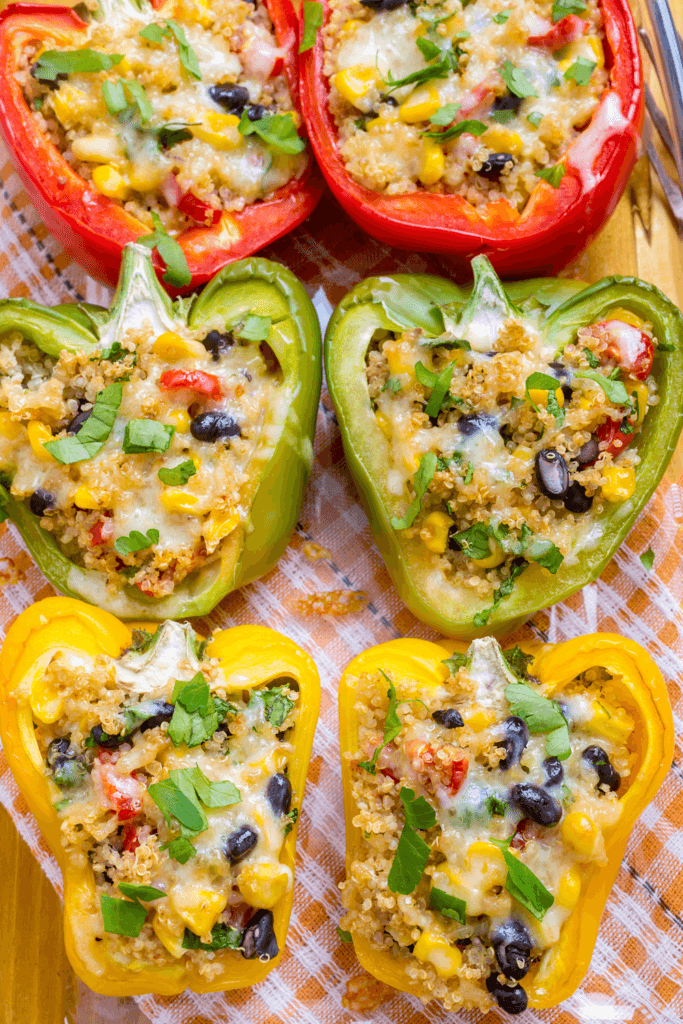 Six colorful bell pepper halves filled with a mixture of quinoa, black beans, corn, chopped vegetables, and melted cheese, garnished with fresh herbs, on a white plate.