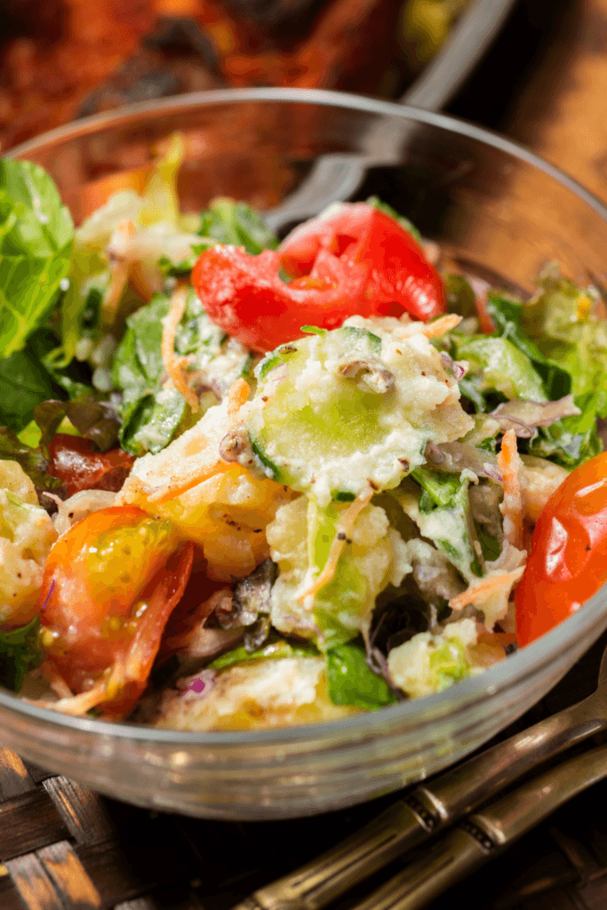 A close-up of a bowl of chicken salad with fennel, cherry tomatoes, cucumber slices, shredded carrots, and leafy greens, all dressed with a creamy yogurt dressing.