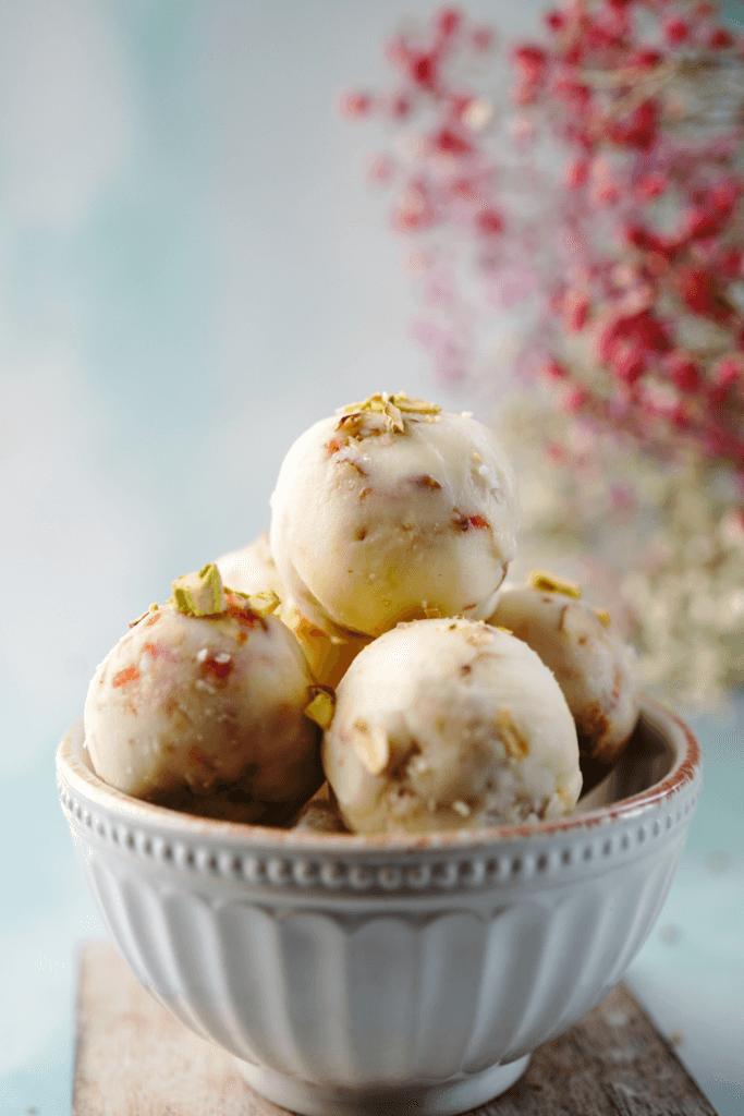 A bowl filled with no-bake cinnamon protein balls, topped with small green pieces of pistachios, placed on a wooden surface with a soft focus background of pink and white flowers.