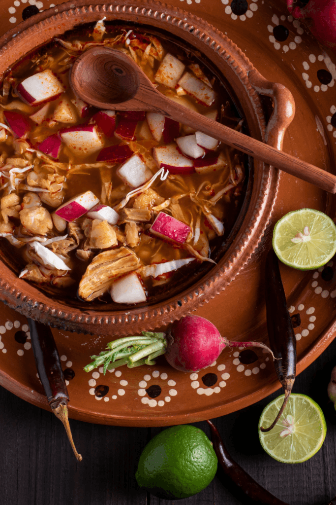 A bowl of traditional Mexican Pozole Rojo, a stew made with pork and hominy, garnished with diced radishes, lime wedges, and chili peppers.