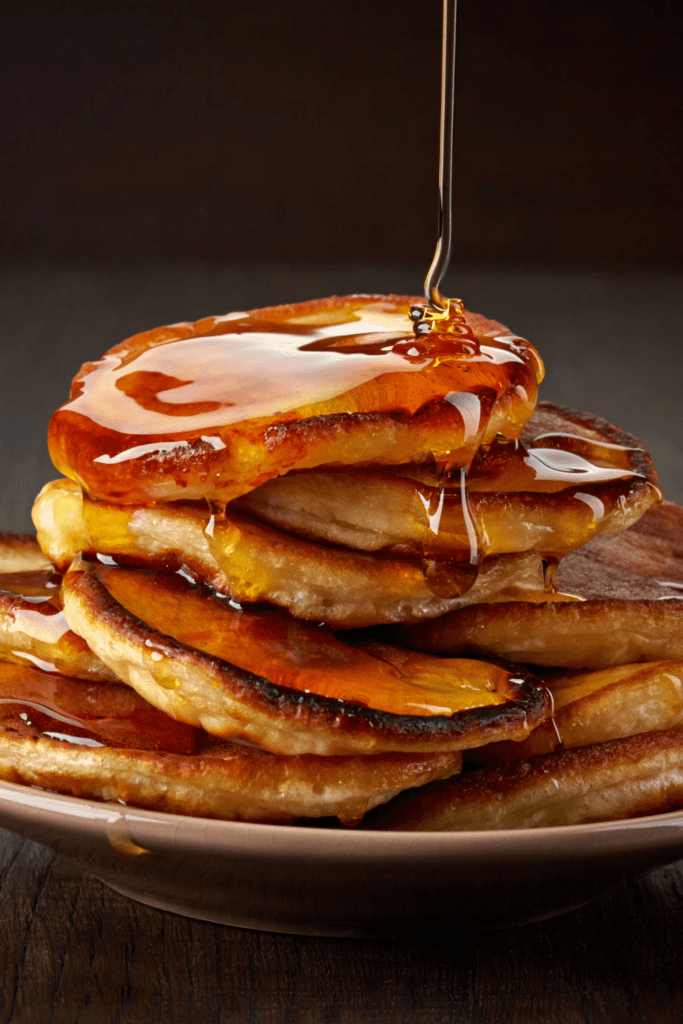 A stack of golden-brown pancakes on a plate with syrup being poured over them, creating a visually appealing and appetizing scene.
