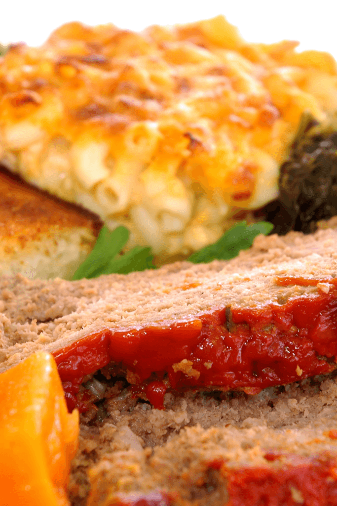A plate of juicy Southern-style meatloaf topped with a red sauce, accompanied by mac and cheese with a golden-brown crust and a portion of sautéed greens.
