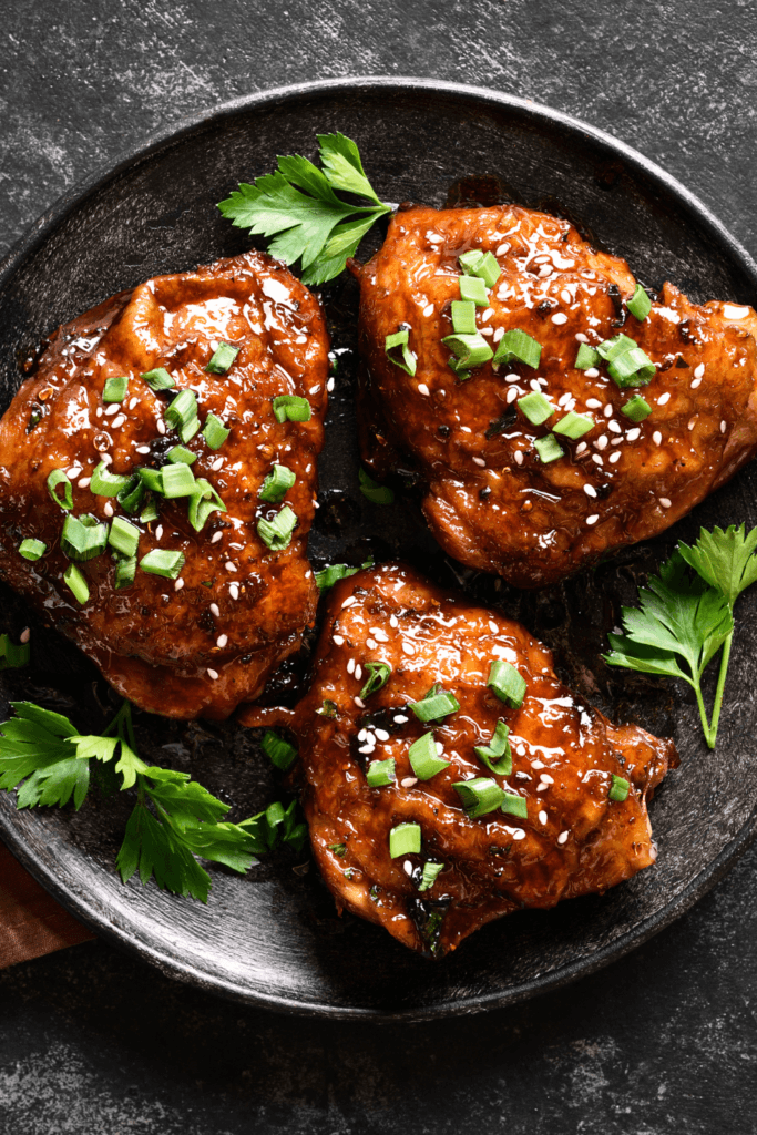 A dish of 5-Ingredient Honey Garlic Chicken with three glazed chicken pieces, garnished with chopped green onions, sesame seeds, and fresh parsley leaves.
