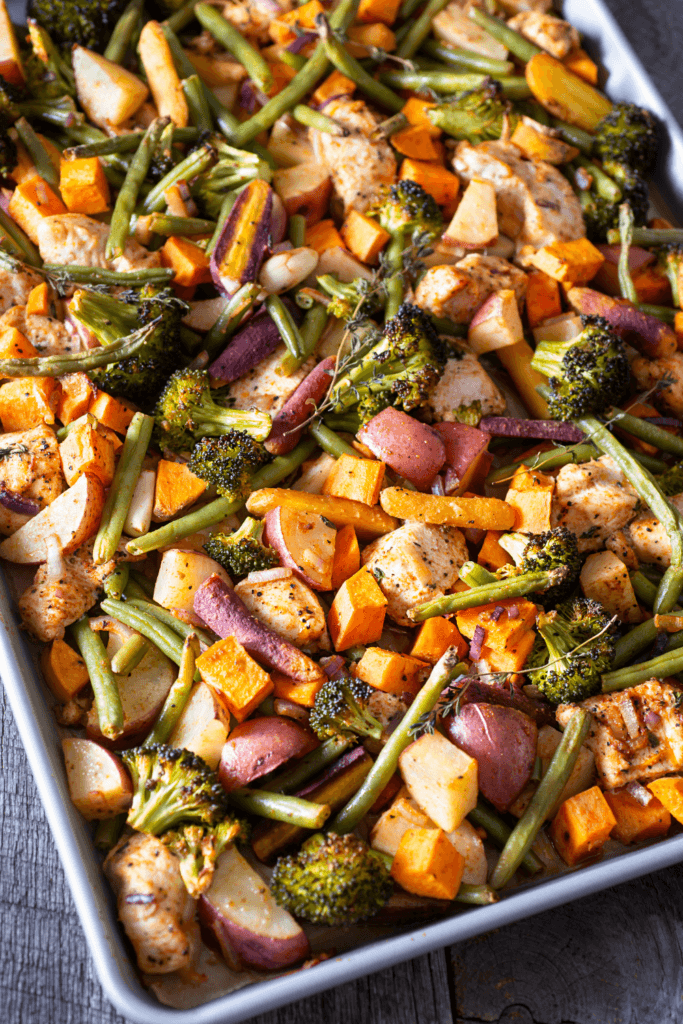 A sheet pan filled with roasted vegetables including green beans, broccoli, sweet potatoes, red potatoes, and carrots, along with seasoned chicken pieces.