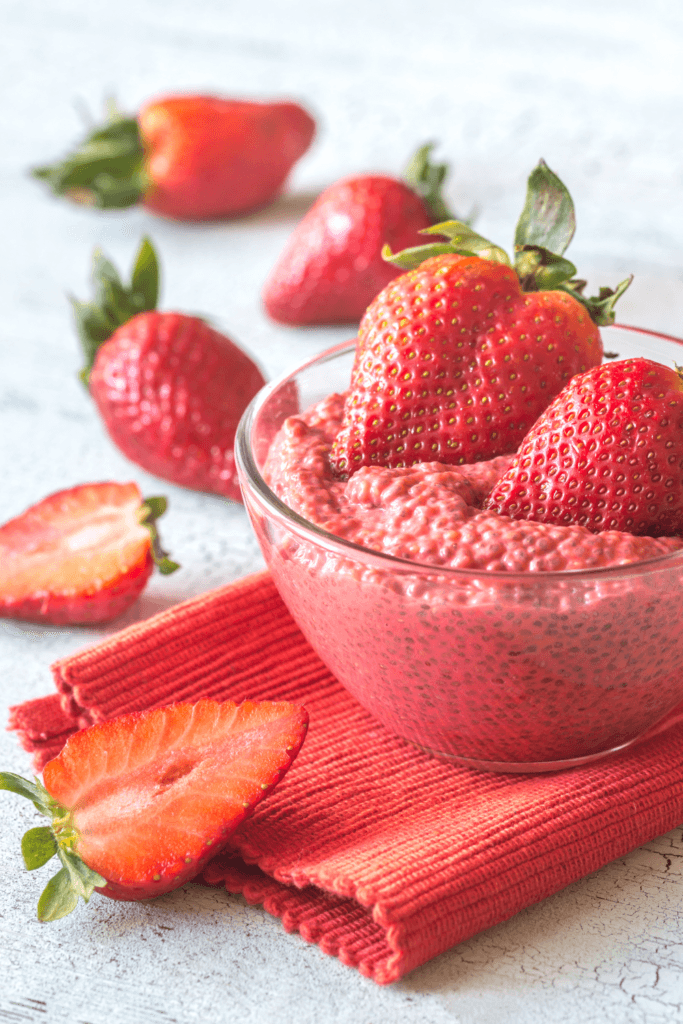 A bowl of strawberry protein pudding garnished with whole strawberries, placed on a red cloth with additional whole and sliced strawberries scattered around.