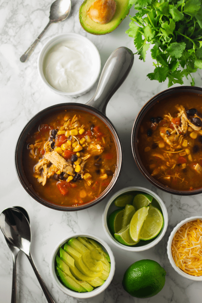 Two bowls of keto chicken tortilla soup with corn, black beans, shredded chicken, tomatoes, avocado slices, sour cream, cheese, and cilantro, surrounded by fresh ingredients.