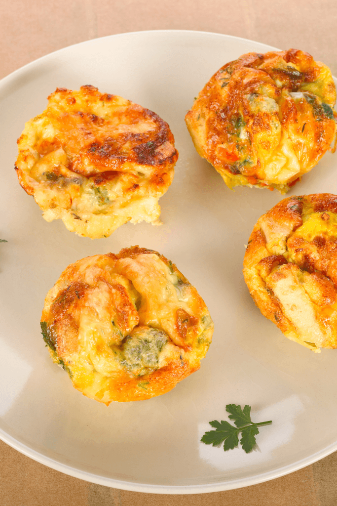 Four egg muffins made with eggs, cheese, and vegetables, such as spinach and bell peppers, cooked in an air fryer and served on a white plate.