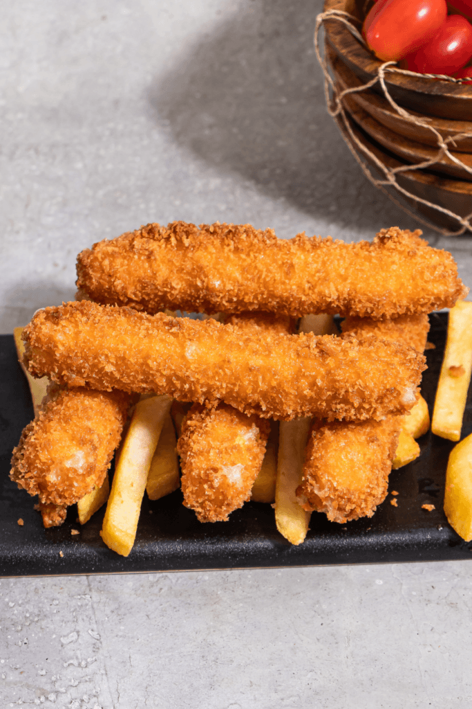 A plate of crispy fried cod fish sticks served with French fries, with a wooden bowl of cherry tomatoes in the background.