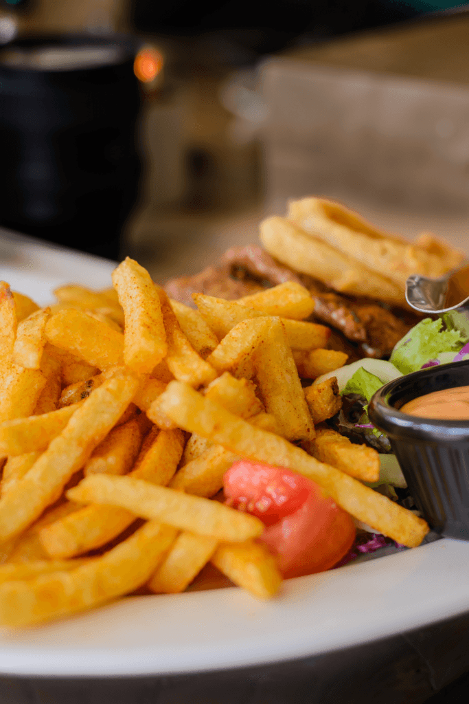 A plate of golden, crispy French fries, onion rings, a small portion of salad with a tomato slice, and a small black container of dipping sauce.