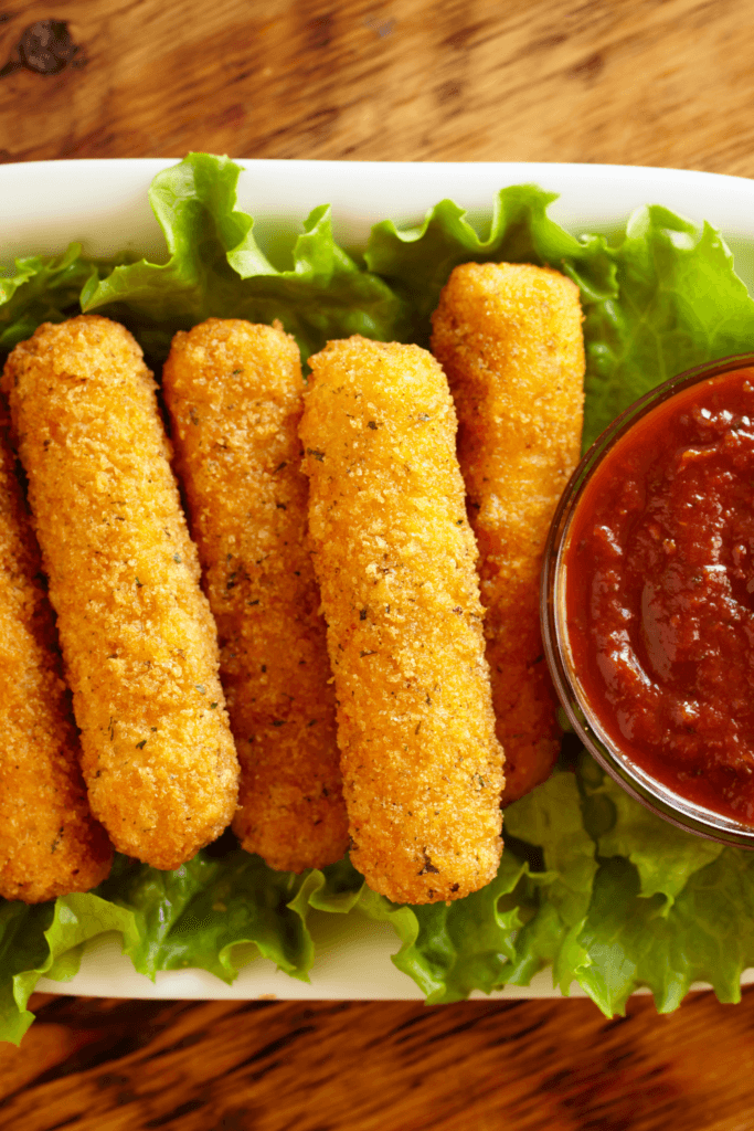 A plate of breaded and fried mozzarella sticks on a bed of green lettuce leaves, served with a small bowl of marinara sauce for dipping.