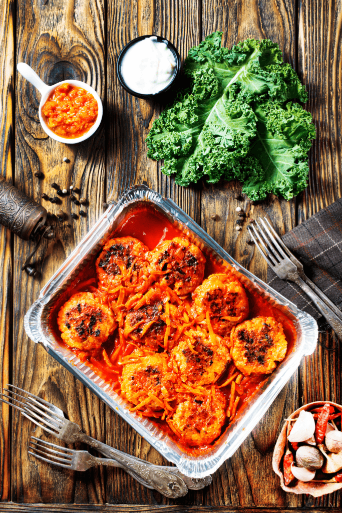 A rectangular aluminum tray filled with BBQ Sriracha ground chicken meatballs topped with shredded carrots, accompanied by a white bowl of orange sauce, a black bowl of creamy sauce, fresh kale leaves, forks, a brown napkin, and a bowl with garlic and red chili peppers, all presented on a wooden table.
