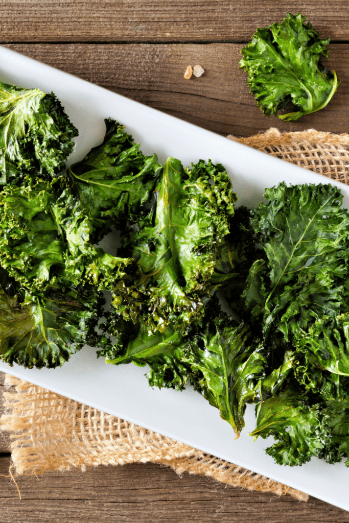 A plate of crispy kale chips cooked in an air fryer, seasoned with pink Himalayan salt, and placed on a white rectangular plate on a wooden surface.