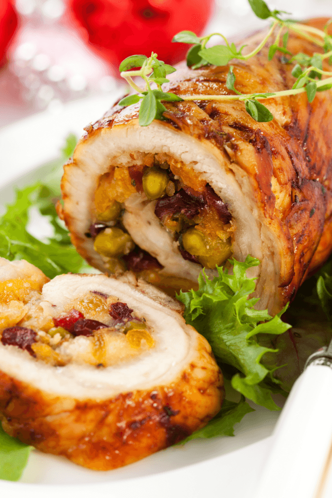 A baked stuffed chicken breast sliced to reveal its filling of pistachios, dried cranberries, and apricots, garnished with fresh herbs and served on a bed of leafy greens.