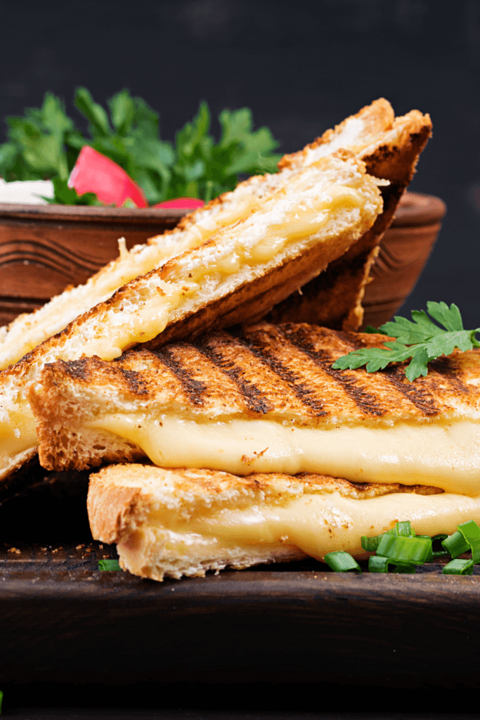 A close-up of a grilled cheese sandwich with grill marks, cut in half to reveal melted cheese inside, with green herbs and a wooden bowl with greens in the background.