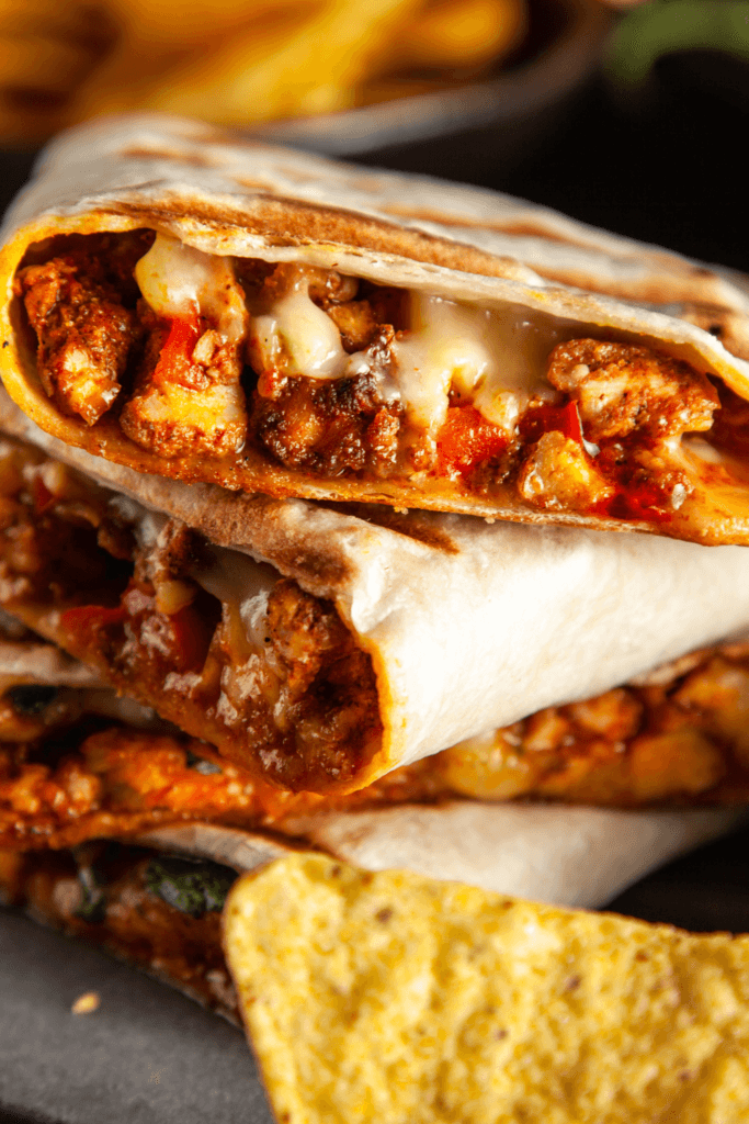A close-up of a grilled quesadilla filled with melted cheese, seasoned meat, and vegetables, cut in half to reveal the filling, with tortilla chips and fries in the background.