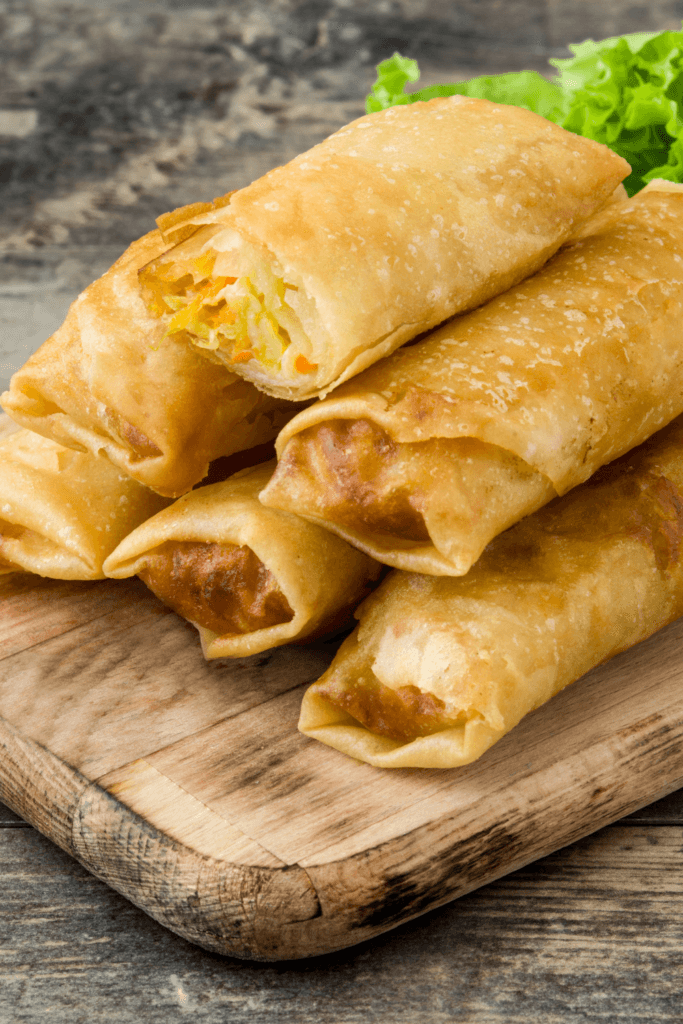 A stack of golden-brown vegetable spring rolls placed on a wooden cutting board, filled with shredded carrots, cabbage, and other vegetables.