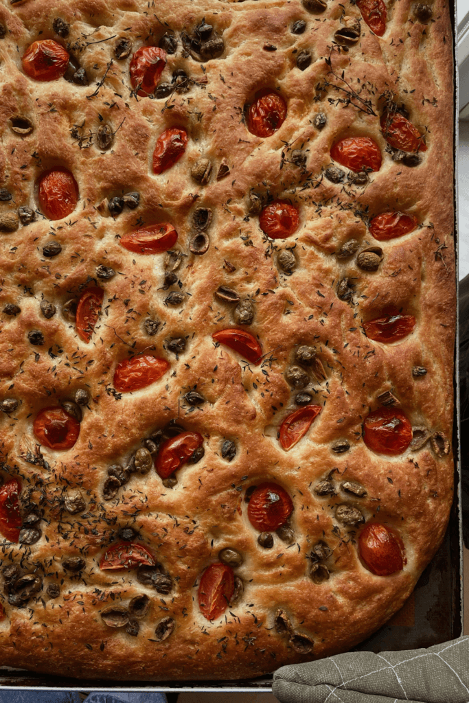Close-up of freshly baked focaccia bread topped with halved cherry tomatoes, olives, and herbs, showcasing a golden-brown crust and characteristic dimples.