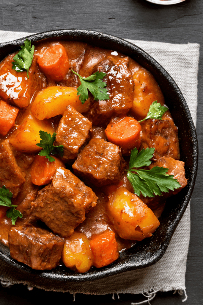Close-up of a hearty beef stew in a black cast iron skillet, featuring chunks of beef, carrots, potatoes, and fresh parsley garnish.