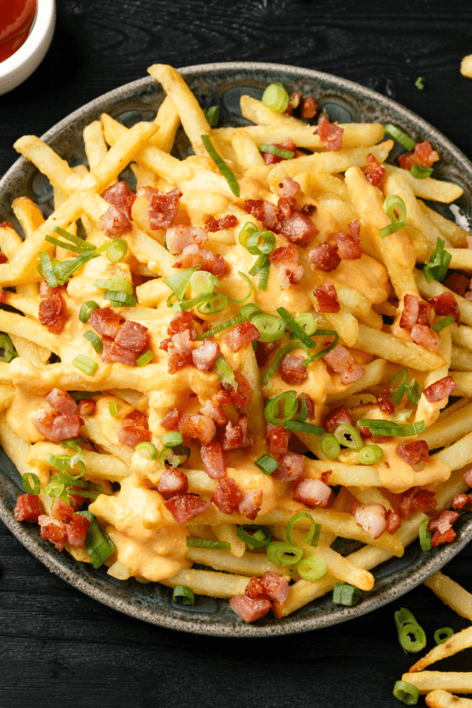 Close-up of loaded French fries topped with melted cheese, crispy bacon bits, and chopped green onions, with a small bowl of ketchup.