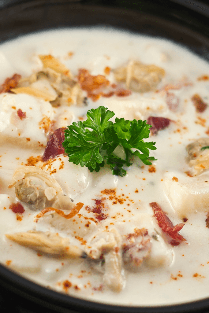 Close-up of a bowl of creamy clam chowder with chunks of clams, pieces of bacon, and a garnish of fresh parsley, seasoned with a sprinkle of paprika.