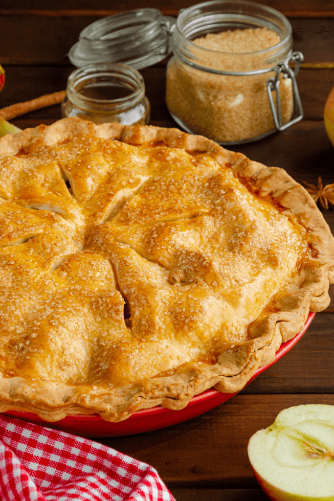Classic homemade apple pie with a golden-brown crust, sprinkled with sugar, served in a red pie dish, with brown sugar, cinnamon, a checkered cloth, and a halved apple visible.