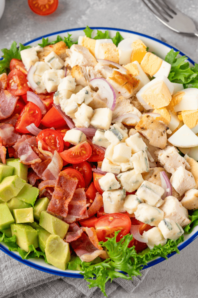 Cobb salad with chopped salad greens, tomatoes, bacon, grilled chicken breast, hard-boiled eggs, avocado, Roquefort cheese, and red-wine vinaigrette, arranged in sections on a white plate.