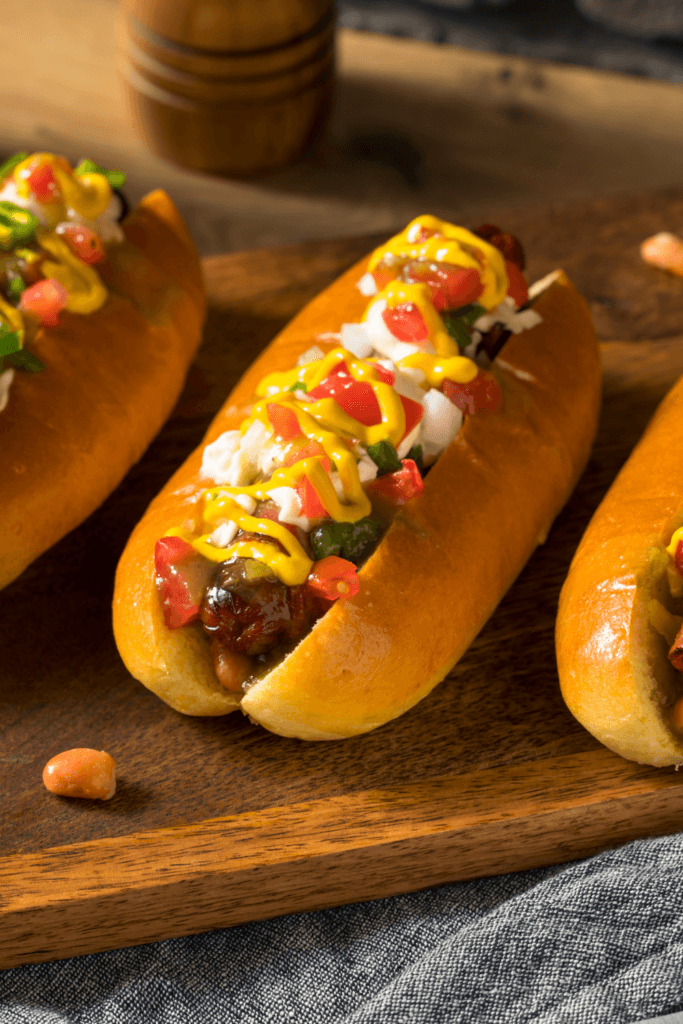 Three hot dogs with mustard, chopped onions, diced tomatoes, and possibly relish, placed on a wooden surface.