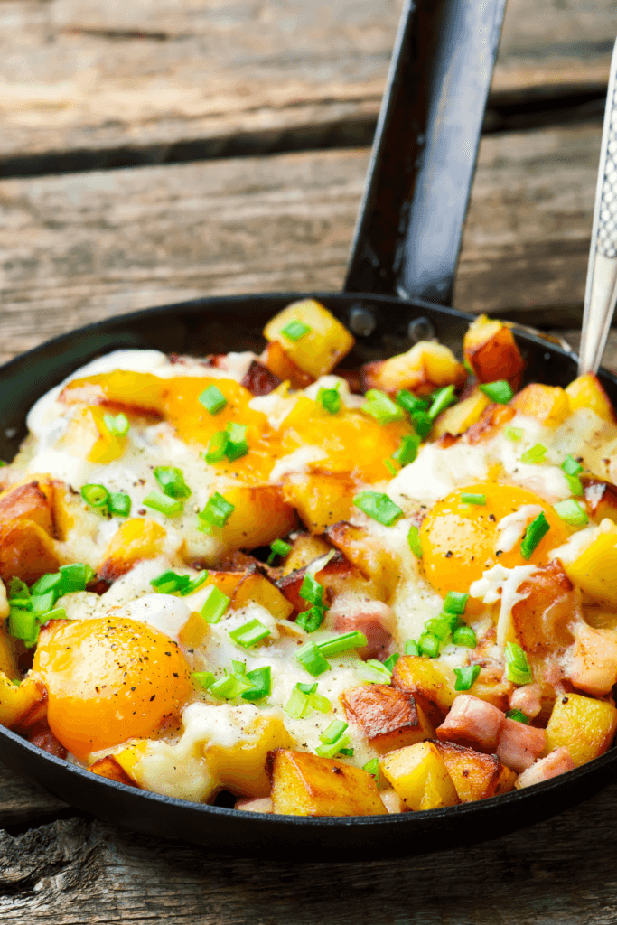 A skillet filled with cheesy bacon and egg hash, featuring golden-brown diced potatoes, crispy bacon pieces, melted cheese, and sunny-side-up eggs, garnished with chopped green onions.