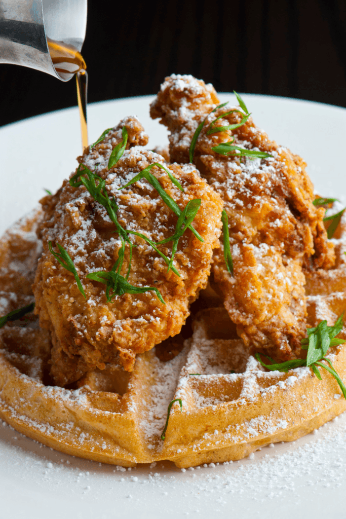 Plate of butter roasted chicken and grit waffles, garnished with green herbs and sprinkled with powdered sugar, with syrup being poured over the dish from a small metal pitcher.
