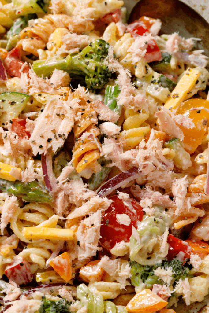 Close-up of a creamy tuna salad with chunks of tuna, broccoli florets, red onion slices, cherry tomatoes, diced bell peppers, and spiral pasta, mixed with a creamy dressing and sprinkled with black pepper.