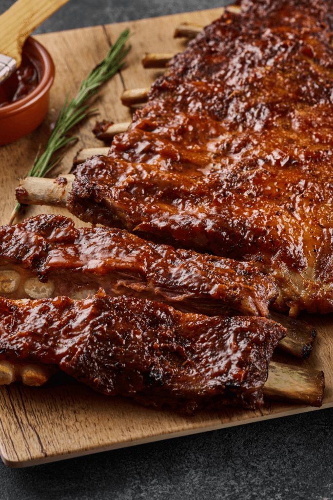 Close-up of sticky BBQ pork ribs on a wooden cutting board, coated with a glossy barbecue sauce, with a sprig of rosemary and a small bowl of additional sauce in the background.