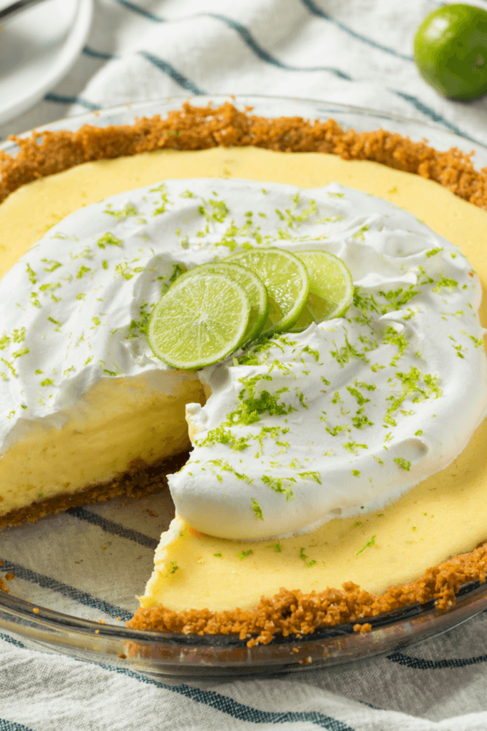 A key lime pie with a slice removed, featuring a graham cracker crust, creamy yellow filling, whipped cream topping, lime slices, and lime zest, displayed in a glass pie dish.