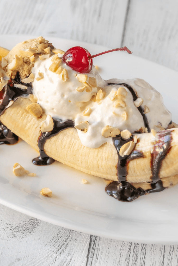 Banana split dessert with vanilla ice cream, chocolate syrup, chopped nuts, and a maraschino cherry, served on a white plate.