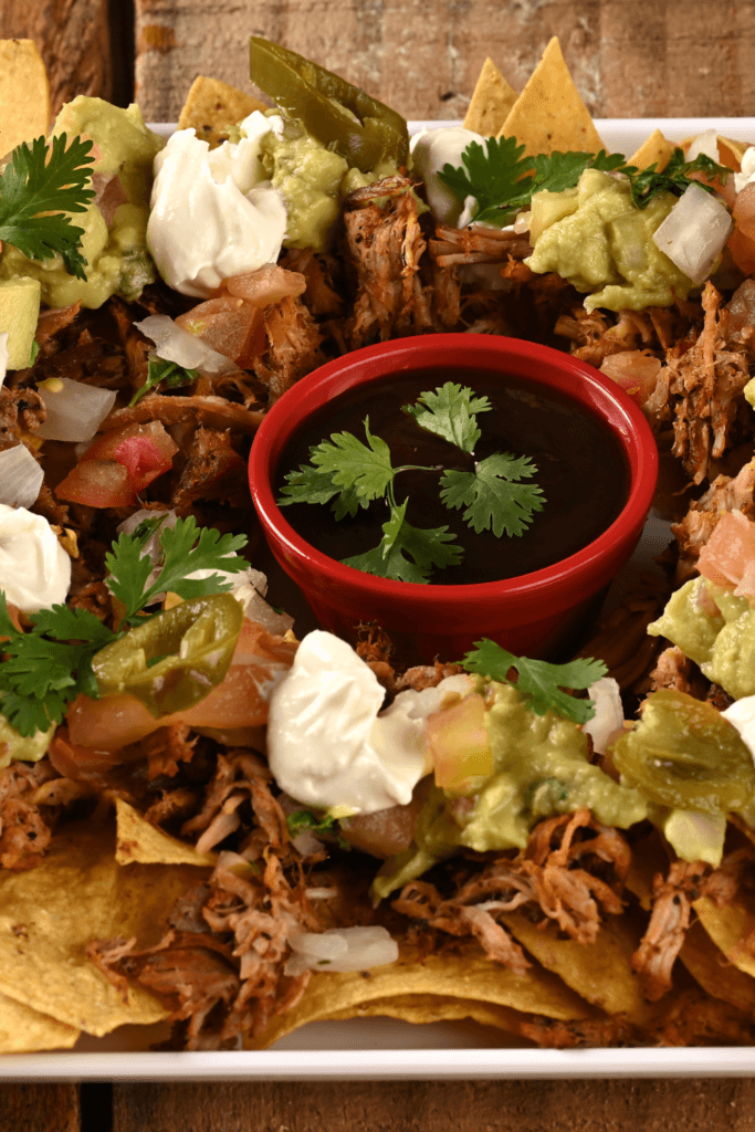 Plate of nachos topped with shredded meat, guacamole, sour cream, diced tomatoes, onions, jalapeño slices, and cilantro, with a bowl of dipping sauce in the center.