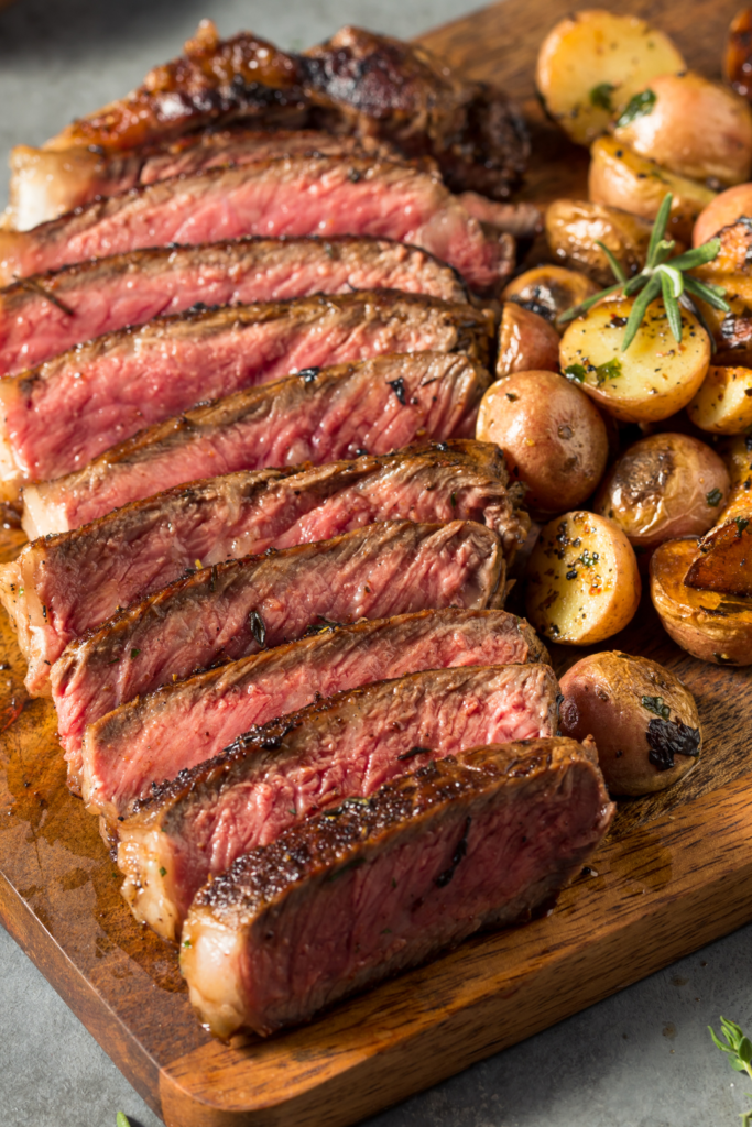 Close-up of a medium-rare steak sliced on a wooden cutting board, served with roasted baby potatoes garnished with herbs.