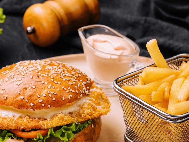 Close-up of a crispy chicken sandwich with lettuce, tomato, and a breaded chicken patty, served with French fries and a small container of dipping sauce.